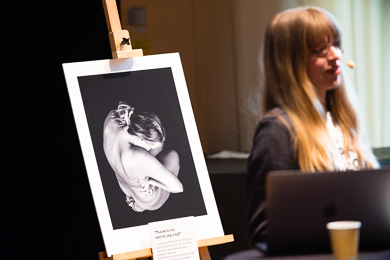 I förgrunden Ulrika "Mohlavyr" Mohlin i oskärpa, i bakgrundens skärpa ett fotografi på staffli från hennes utställning "Kära kropp"